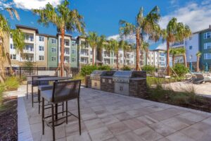 Modern apartment courtyard with palm trees, outdoor grills, and seating area under a clear blue sky.