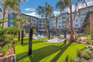 Modern apartment complex with palm trees and hammocks beside a pool under a sunny blue sky.
