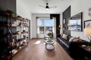 Modern living room with black sofa, bookshelf, ceiling fan, and natural light from window. Cozy, minimalist decor.