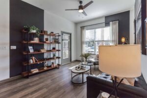 Modern living room with dark accent wall, wood shelving, ceiling fan, and cozy furniture. Bright, natural light filters in.