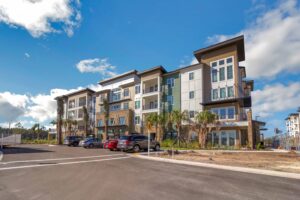 Modern apartment building with blue sky background, palm trees, and parked cars in the foreground.