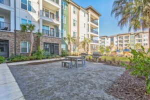 Modern apartment complex with outdoor seating, grills, palm trees, and landscaped greenery under a clear blue sky.