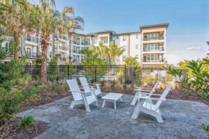 Modern apartment building with palm trees and outdoor seating area in front. Sunny day scenery.