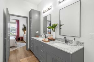 Modern bathroom with gray double vanity, granite countertop, large mirrors, and view into a bedroom with pink curtains.