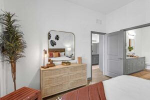 Modern bedroom interior with cozy bed, wooden dresser, mirror, and decorative plants. Soft lighting creates a warm ambiance.
