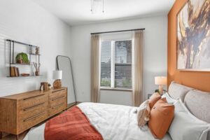 Modern bedroom with orange accent wall, cozy bedding, wooden dresser, large window, and stylish decor.