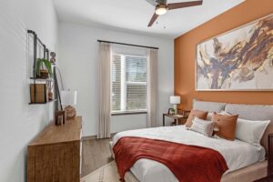 Modern bedroom with warm orange accents, abstract wall art, and natural light from a window with beige curtains.