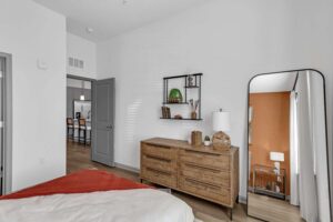Modern bedroom with wooden dresser, large mirror, and wall decor. Cozy, minimalist design with orange accent wall.