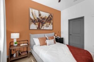 Modern bedroom with orange accent wall, abstract painting, cozy bed, and stylish decor elements.